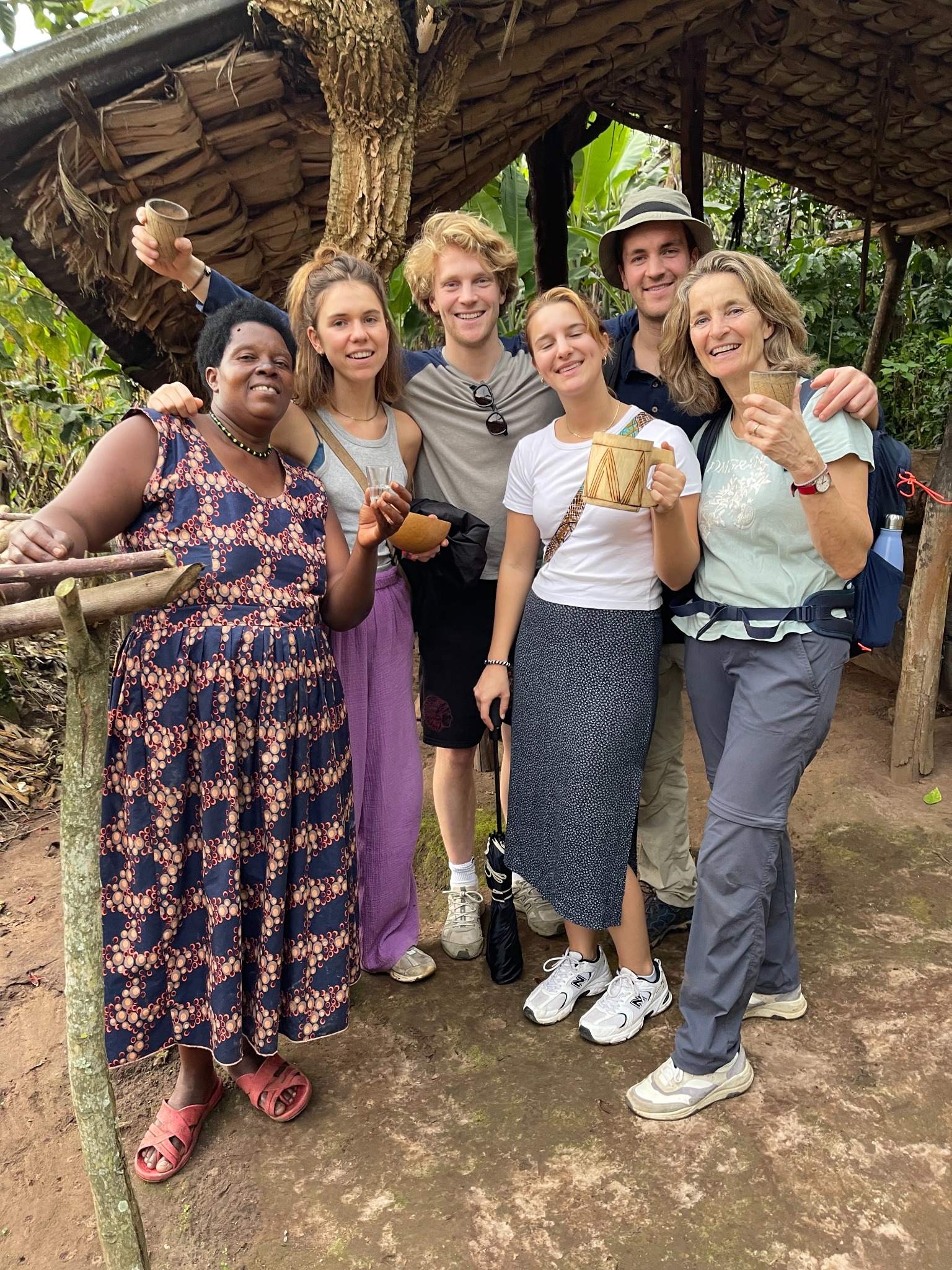 Six guests and a woman in a printed dress raising cups under a thatched shelter, forest behind