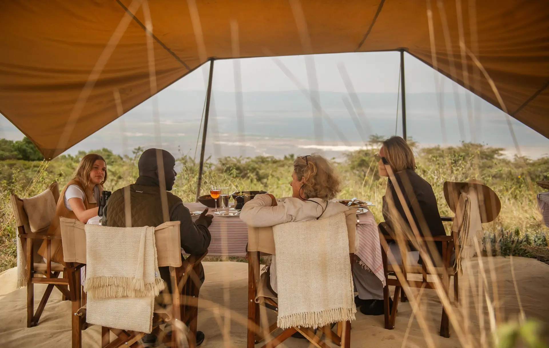 Four guests at a long table under a canvas awning, a woman standing beside them, a plain and lake beyond